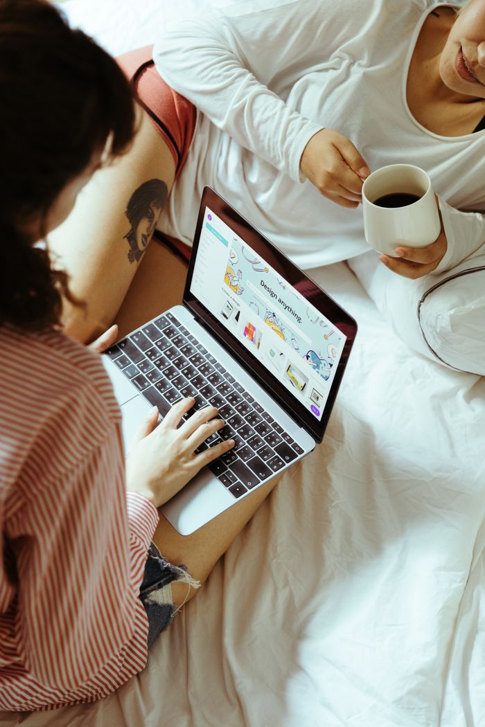 From above anonymous women couple lesbian resting on bed and browsing laptop while spending time together and drinking coffee during weekend