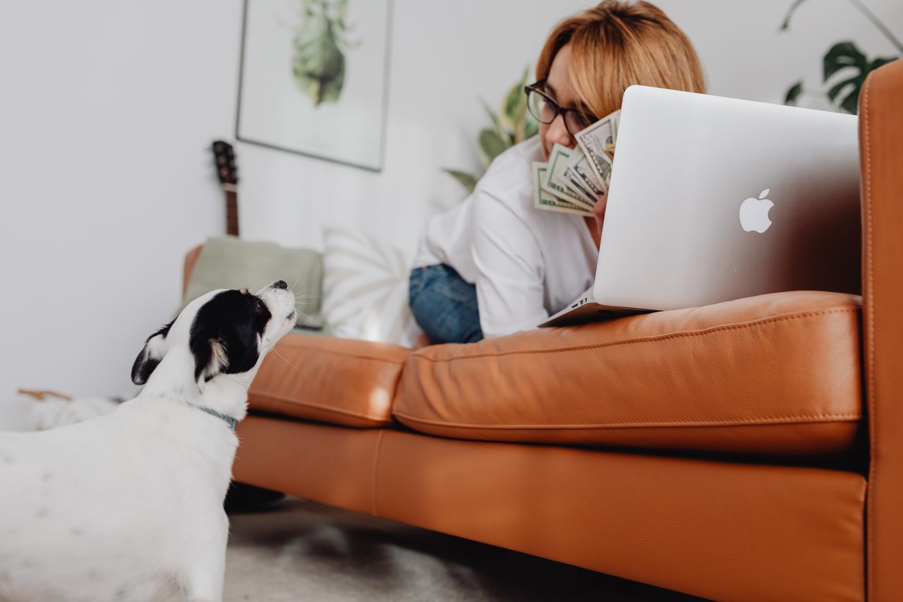 Woman lying on orange sofa with dog, laptop, and dollar bills, creating a cozy living room scene.